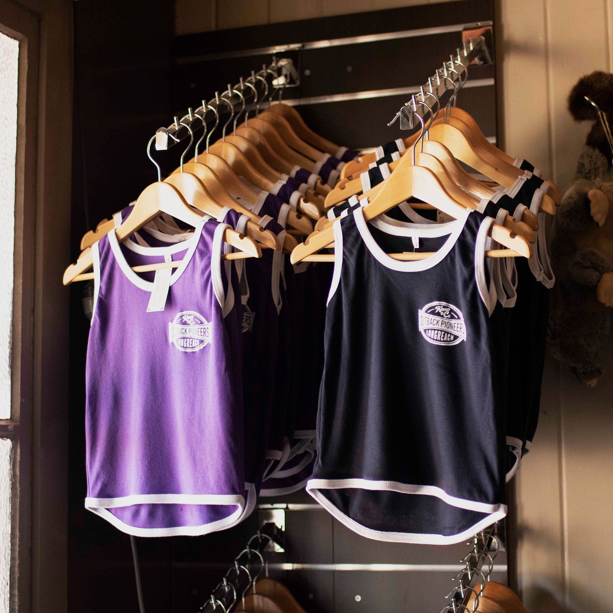 Two shearing singlets side by side on wooden coat hangers. One is purple and one is navy blue.