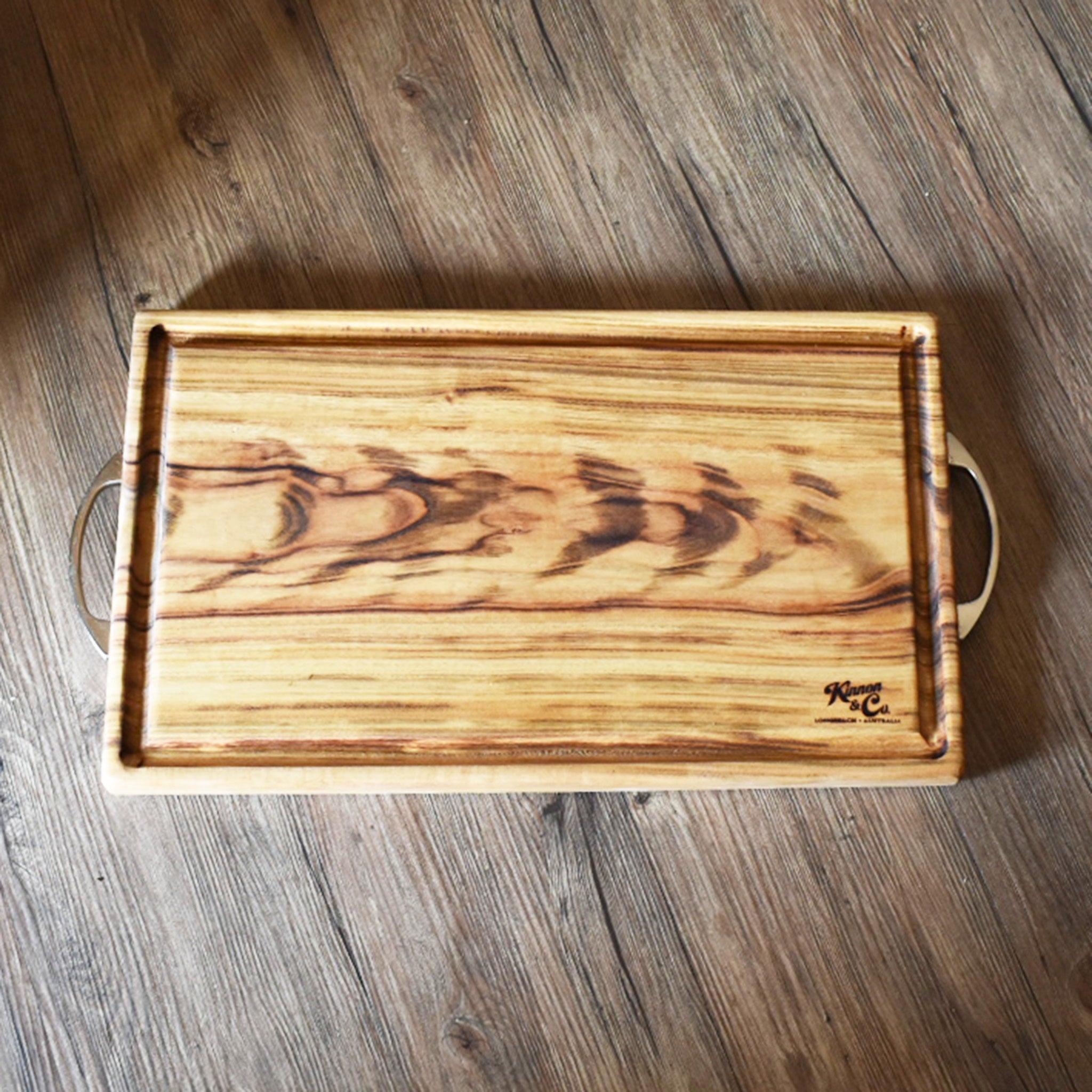 Large timber cutting board with two silver handles , one at either end. Shown on rustic wooden background.