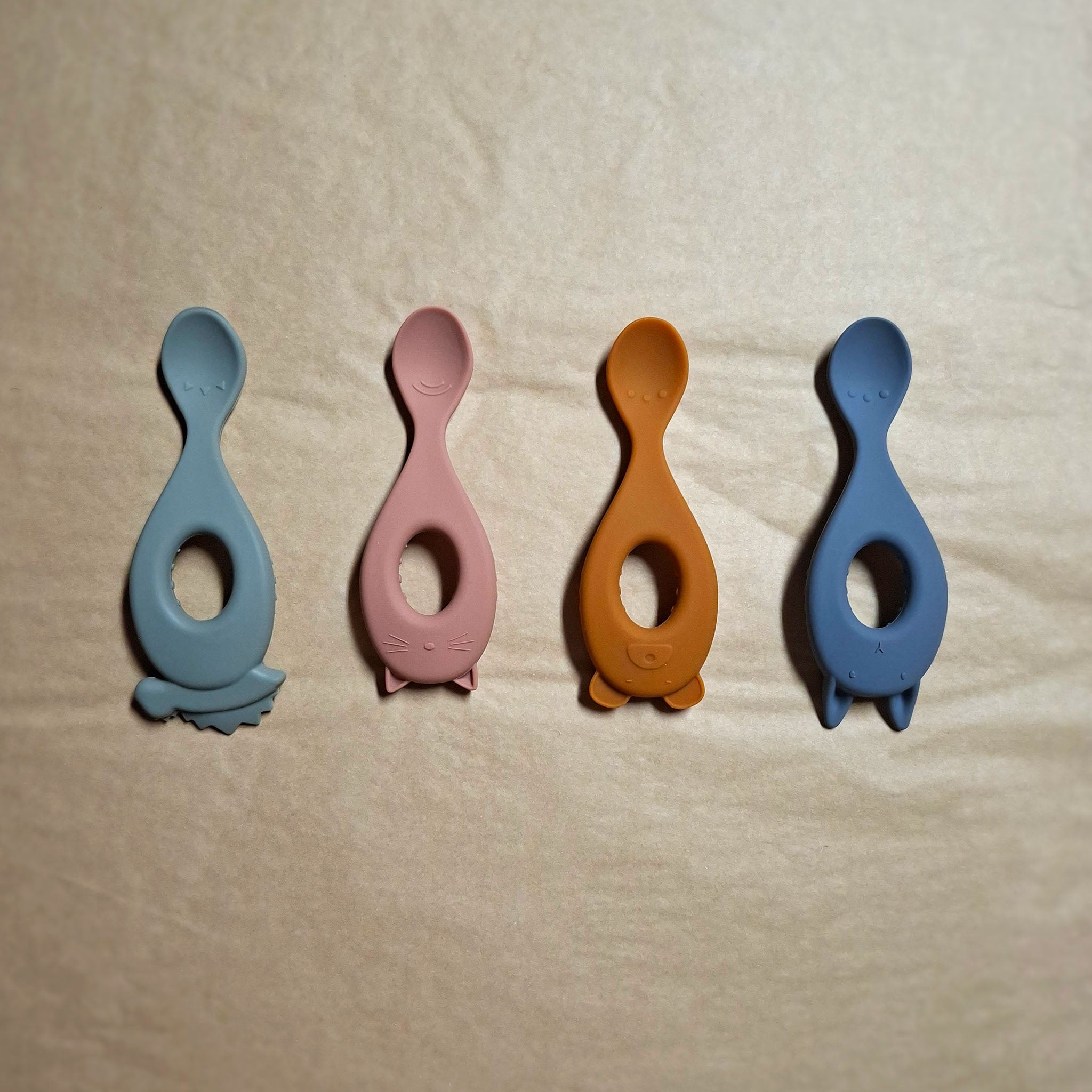 Four silicone baby feeding spoons in green, yellow, blue, pink on a beige background